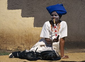 SOUTH AFRICA, Lesedi, Xhosa lady smoking pipe on street with head wear and painted face.