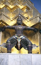 THAILAND, Bangkok, Wat Phra Kaew, Or Grand Palace. Detail of statue standing against golden wall