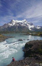 CHILE, Patagonia, Torres Del Paine, Man sitting on the riverbank beside rapids below the distant