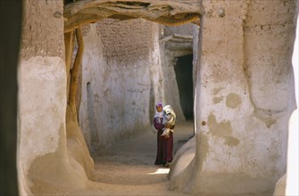 EGYPT, Western Desert, Dhalla Oasis, Woman carrying child along street between mud brick buildings.