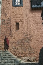 TIBET, Xigatse, Tashilumbo Monastery, Monk standing on steps in front of peach coloured painted