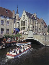 BELGIUM, West Flanders, Bruges, Tourists on boat trips on a canal with old bridge and houses behind