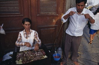 BOLIVIA, Tarija, Women cooking meat skewers on barbecue with man eating nearby