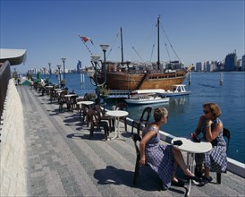 UAE, Abu Dhabi, Corniche. Restaurant Dhow by seam moored boats with two woman sat on table and