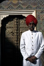 INDIA, Rajasthan, Jaipur, Palace guard dressed in white and wearing a red turban standing by ornate