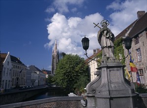 BELGIUM, West Flanders, Bruges, Religious statue on bridge over canal with church spire in distance