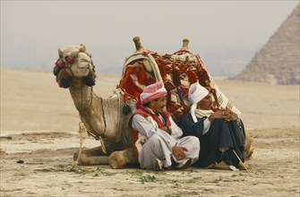 EGYPT, Cairo Area, Giza, Camel lying down with two men seated on one side. Pyramid in background