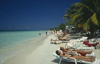 WEST INDIES, Jamaica , Negril, Sunbathers on white loungers on sandy beach lined by palm trees.