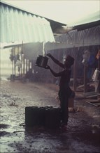 CAMBODIA, Thmar Pouk, Man collecting rainwater as it runs off a roof above him.