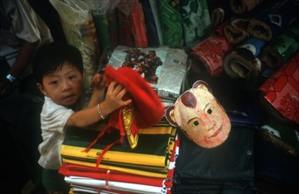 CAMBODIA, Phnom Penh, Central Market.  Small boy in shop selling fabric and flags. Coloured mask