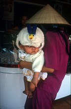 VIETNAM, Tay Ninh, Woman in a straw hat holding a baby as she looks at a shop display in Tay Ninh
