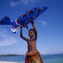 INDONESIA, Lombok, Timur, Young boy in red floral sarong holds up a blue eagle shaped kite