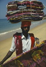 INDIA, Kerala, Kovalam Beach, Fabric salesman on beach carrying stack of colourful materials on his