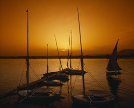 EGYPT, Nile Valley, Aswan, Feluccas at sail and moored at jetty silhouetted against golden sunset.