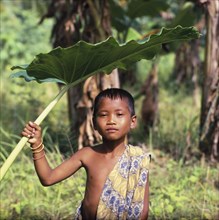 INDONESIA, Lombok, Kuta, Boy with a sarong over his shoulder using a large leaf as sun shade