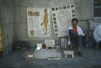 CHINA, Hainan Island, Sanya, Roadside medicine seller seated in front of medical drawings of the