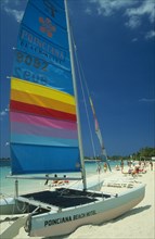 WEST INDIES, Jamaica, Negril, Catamaran with sail up on beach with tourists sunbathing and walking
