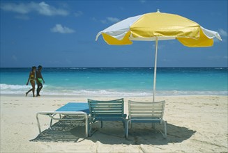 BERMUDA, Long Bay Beach, Tourist couple walking on beach by water past umbrella and sun loungers