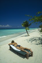 WEST INDIES, Jamaica, Negril, Woman Sunbathing on lounger on beach near sea and coconut palm tree