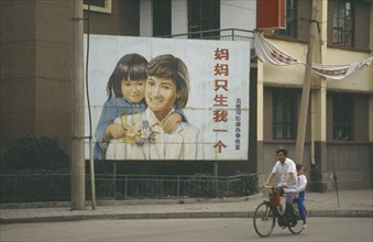 CHINA, Shandong, Jinan, Man with child passenger cycling past birth control poster with image of
