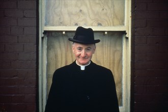 IRELAND,  , Dublin, Priest wearing hat in front of boarded up house window.