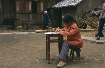 CHINA, Guizhou, Young girl doing homework at desk in farmyard.
