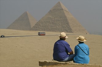 EGYPT, Cairo Area, Giza, The Pyramids with two tourists seated in foreground. Bus on road.