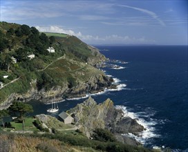 ENGLAND, Cornwall, South Cornwall . Rugged coastline with yachts in bay and houses on hillside