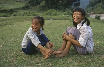 CHINA, Guangxi, Two farm girls sitting on grass laughing.