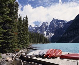 CANADA, Alberta, Moraine Lake, Canoes on a jetty beside the pine tree lined lake with snow caped