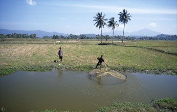 MALAYSIA, Kedah, Langkawi, Two men by pond in fields near Cenang with one casting a fishing net