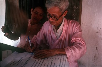 VIETNAM, Religion, Seated Buddhist Monk writing whilst being fanned by a woman on his right.