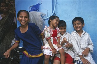INDIA, Maharashtra, Mumbai, Four smiling children leaning against bicycle with blue wall behind.