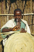 ETHIOPIA, Harerge Province, "Woman making circular mat from straw, stitched into place."