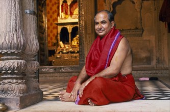 INDIA, Utter Pradesh, Vrindaven, Hindu priest in red robes sitting cross legged on temple floor.
