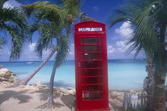 WEST INDIES, Antigua, Dickenson Bay, Red telephone box on sand surrounded by palm trees with sea