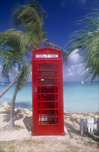 WEST INDIES, Antigua, Dickenson Bay, Red telephone box on sand surrounded by palm trees with sea
