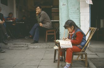 CHINA, Sichuan, Leshan, Young girl doing homework at small table outside house watched by adults in