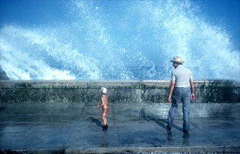 CUBA, Havana, Malecon, Old man and small boy standing on pavement with waves crashing over the sea