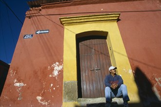 MEXICO, Oaxaca, Oaxaca City, Man in baseball hat sitting on stone step of doorway framed by yellow