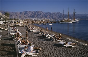 ISRAEL, Eilat, Coral Beach, "Lines of sunbathers on white loungers on dark, sand beach with sailing