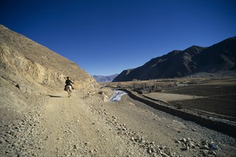 CHINA, Tibet, Shigatse, Man on pony riding along dusty road leading past ploughed fields