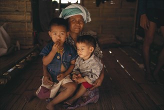 THAILAND, North, Mae Sai Area, Karen Refugee mother sitting with her two children on the floor of a