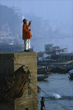 INDIA, Uttar Pradesh, Varanasi, "Holy man praying on stone wall above River Ganges,boats,town,mist