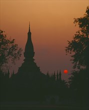 LAOS, Vientiane, Pha That Luang Sacred Stupa silhouetted against red setting sun and orange sky.