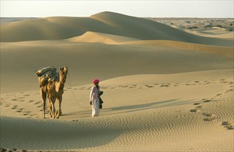 INDIA, Rajasthan, Jaisalmer, Thar Desert.  Man walking with camel carrying pack over dunes leaving