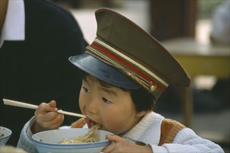 CHINA, North, Children, Boy wearing peaked military cap eating noodles from bowl with chopsticks.
