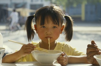 CHINA, Guizhou, Guiyang, Pony tailed young girl eating noodles from bowl with chopsticks