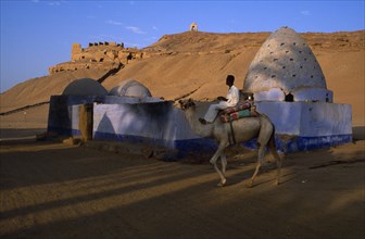 EGYPT, Upper Egypt, Aswan, Man on camel rides past Beehive Mausoleum with Tomb of the Nobles on