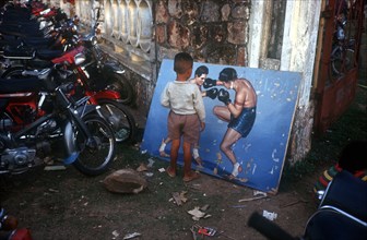 CAMBODIA, Battambang, Small boy looking at painting of boxers propped against the wall in a